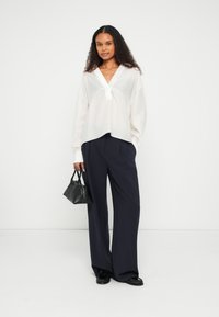 Løs, hvit skjorte med v-hals og plissédetalj, kombinert med mørkeblå vide bukser og en liten svart håndveske.