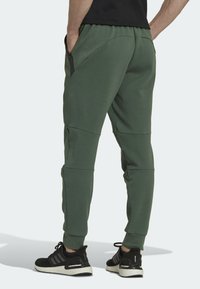 Zelené sportovní kalhoty s zužujícím se designem, elastickým pasem, bočními kapsami a kontrastními černými zipovými akcenty. V kombinaci s černými teniskami.