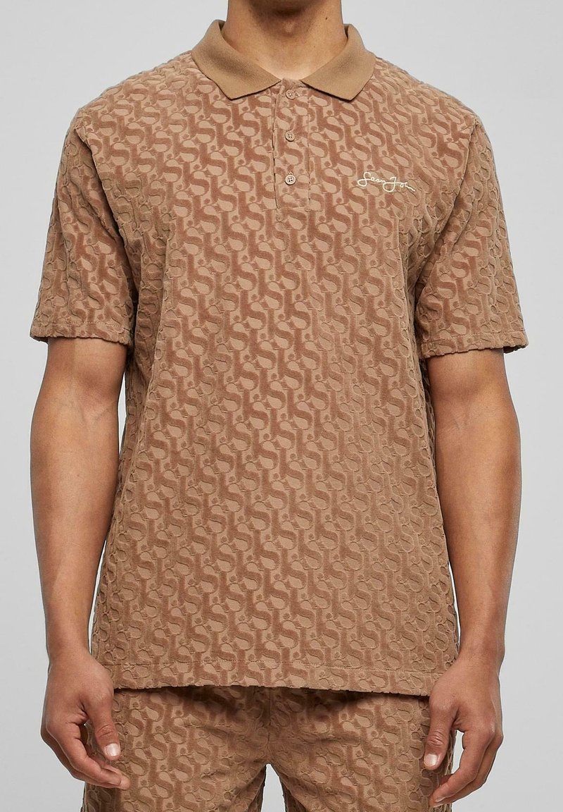 Homme portant un polo marron texturé et un short assorti avec un motif abstrait répétitif et un logo blanc brodé sur la poitrine.