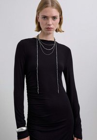 Vestido negro de manga larga con textura fruncida en un lado. La modelo lleva collares plateados en capas y una pulsera plateada con textura.