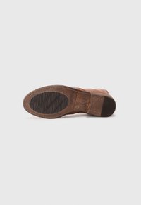 Semelle de botte en cuir avec un dessus texturé marron, présentant un bout courbé et un talon légèrement surélevé. Détails cousus et motifs de grip en caoutchouc.