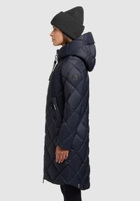 Tumes sinine tikitud puffer jakk, millel on kapuuts. Omab lukuga sulgemist, küljetaskuid ja ümmargust logoaplikatsiooni vasakul varrukal. Kootud hall beanie.
