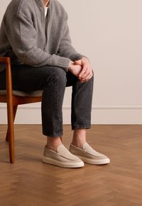 Beige Slipper mit einem glatten Textilobermaterial und weißer Gummisohle, kombiniert mit dunklen Jeans und einer grauen Strickjacke.