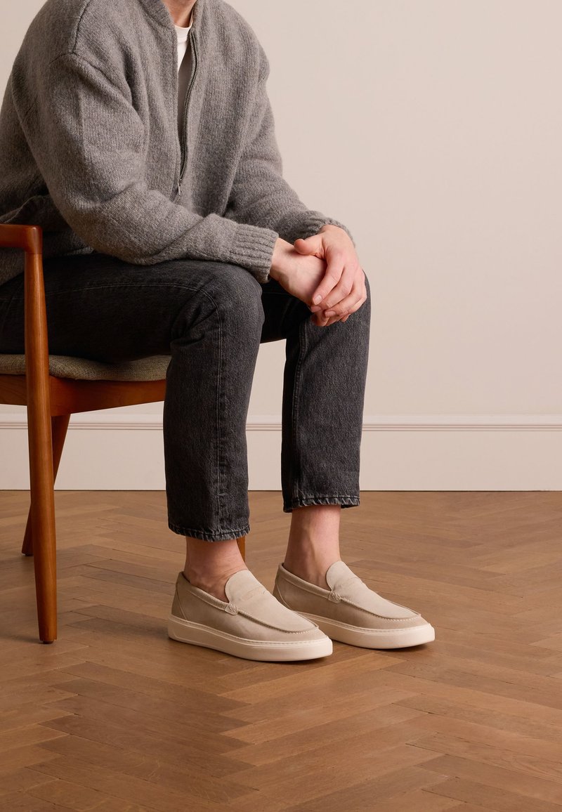 Beige Slipper mit einem glatten Textilobermaterial und weißer Gummisohle, kombiniert mit dunklen Jeans und einer grauen Strickjacke.