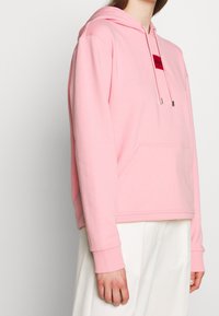 Sweat à capuche rose en tissu doux avec une poche kangourou à l'avant et des cordons de serrage réglables avec embouts métalliques. Porte un patch logo sur la poitrine.