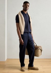Marineblå polo skjorte med korte ermer, beige strikket vest, marineblå bukser og lysebrune semskede loafers. Bærer en stor beige duffelbag.