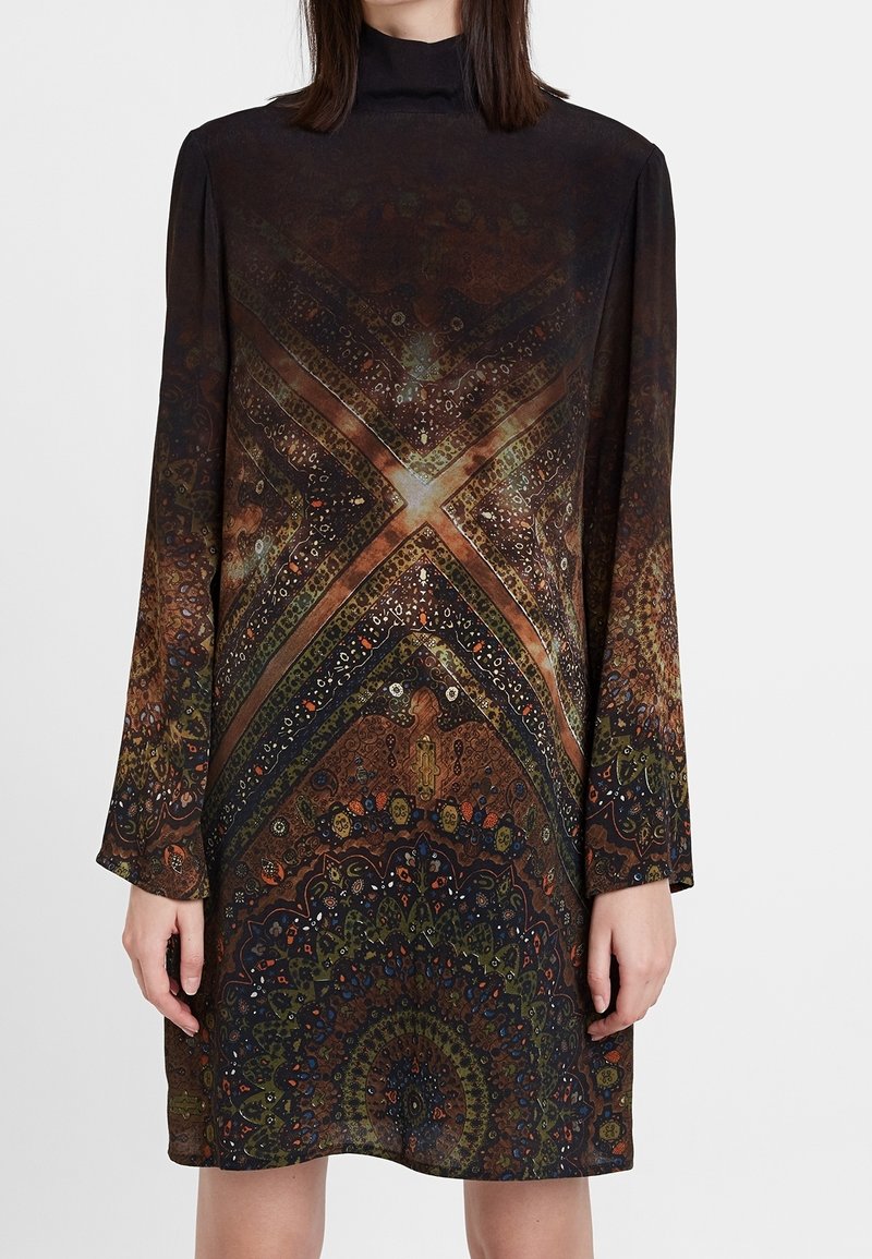 Femme portant une robe à manches longues avec un col haut et des motifs géométriques et floraux complexes en brun foncé, vert et orange.