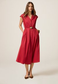 Rotes, kurzärmeliges, knopfbares Kleid mit tailliertem Gürtel, schwingendem Rock und Taschen. Schuhe mit Leopardenmuster ergänzen das Outfit.
