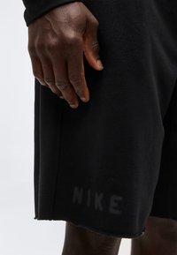 Κοντινό πλάνο ενός σκουρόχρωμου χεριού που ακουμπά κοντά στο τελείωμα μαύρων σορτς με σβησμένο λογότυπο "NIKE".