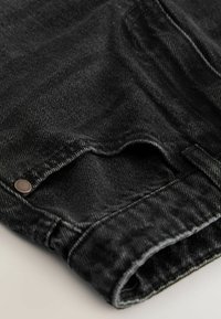 Nahaufnahme von schwarzem Denimstoff, der detaillierte Nähte, einen Metallniet und einen gefalteten Rand eines Kleidungsstücks zeigt.