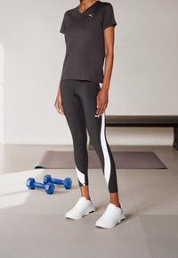 Černé tričko s výstřihem do V, černé legíny s bílými bočními panely a bílé sportovní boty. Na tričku a legínách je malé logo.