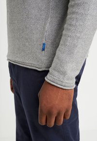 Helles graues Langarmoberteil mit strukturiertem Strick, Rundhalsausschnitt und einem kleinen blauen Markenetikett an der Seite, kombiniert mit dunklen Hosen.