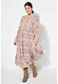 Maxi-jurk met een kleurrijk geometrisch patroon in oranje, paars en teal, met lange mouwen en een gelaagde rok, gedragen met hoge bruine laarzen.