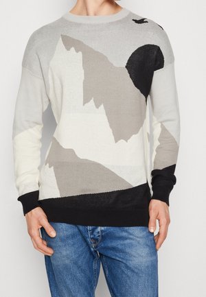 Man draagt een trui met ronde hals met abstracte berg- en zonpatronen in zwart, grijs en wit, gecombineerd met een blauwe spijkerbroek.