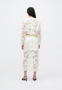 Crèmekleurige kanten jurk met bloemenpatroon, lange mouwen, cropped top en een aansluitende midi-rok. Gecombineerd met bruine sandalen met hak.