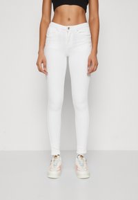 Bele skinny kavbojke v beli barvi, narejene iz denima, s srednjim pasom, petimi žepi in zadrgo z srebrnim gumbom. Nosi se jih s supergami.