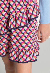 Saia colorida com padrão geométrico e bainha rendada. Apresenta acentos em laranja, rosa e azul-marinho, finalizada com um acabamento decorativo em azul-marinho.