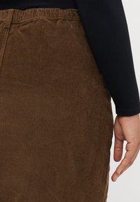 Fustă din corduroy maronie cu o textură ribbed și o talie elastică. O mână se află aproape de marginea fustei, purtând o bluză neagră cu mâneci lungi deasupra.