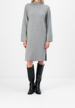 Personne portant une robe-pull grise à manches longues avec des fentes sur les côtés, associée à des bottes en cuir noir montant jusqu'aux genoux, debout devant un fond blanc uni.