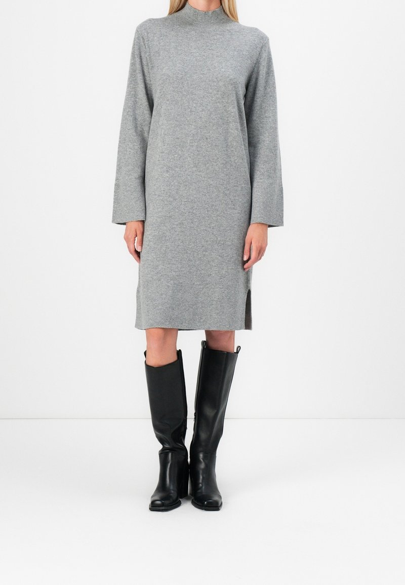 Personne portant une robe-pull grise à manches longues avec des fentes sur les côtés, associée à des bottes en cuir noir montant jusqu'aux genoux, debout devant un fond blanc uni.