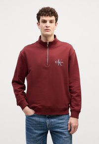 Μπορντό φούτερ με ψηλό λαιμό και φερμουάρ, με λογότυπο στο στήθος. Το υλικό φαίνεται μαλακό, με ριμπ μανσέτες και στρίφωμα.