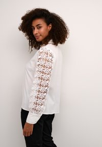 White long-sleeve blouse featuring intricate lace detailing on the sleeves. Soft fabric with a relaxed fit, paired with black trousers.