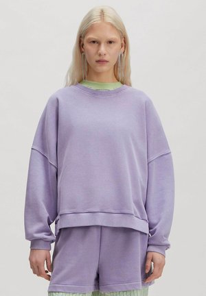 Blonde Person trägt übergroßen lavendelfarbenen Pullover und passende Shorts, ergänzt durch lange hängende silberne Ohrringe, neutraler Gesichtsausdruck.