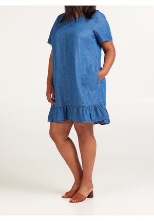 Femme portant une robe en jean bleue à manches courtes avec ourlet volanté et poche ronde, assortie à des sandales marron tressées à talons, debout de manière détendue.