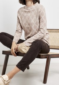 Maglione beige a motivo lavorato a maglia con maniche lunghe, indossato con pantaloni aderenti marrone scuro e mocassini beige, seduto su una sedia in legno con seduta in canna.