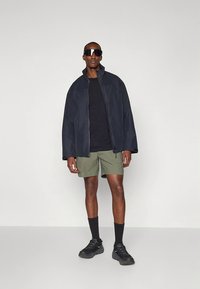 Marinblå zip-up jacka över svart t-shirt, olivgröna shorts, svarta strumpor och skor. Accessoarer inkluderar svarta solglasögon. Släta texturer.