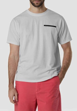 Uomo che indossa una maglietta bianca semplice a maniche corte con piccolo testo nero vicino al petto e pantaloncini rossi vivaci, in piedi con una mano in tasca.