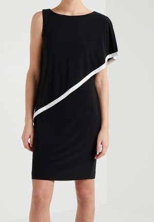 Femme portant une robe noire sans manches avec une bande diagonale blanche asymétrique et une manche drapée sur une épaule.