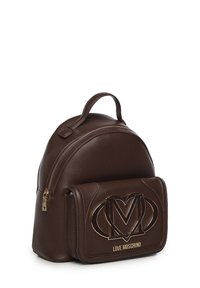 Mochila de imitación de cuero marrón con forma redondeada. Cuenta con un bolsillo frontal con un logo "M" en tono dorado y cierre con cremallera en el compartimento principal.