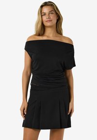 Kobieta z długimi blond włosami, ubrana w czarny top na ramionach i czarną plisowaną spódnicę, stojąca na tle jednolitego białego tła.