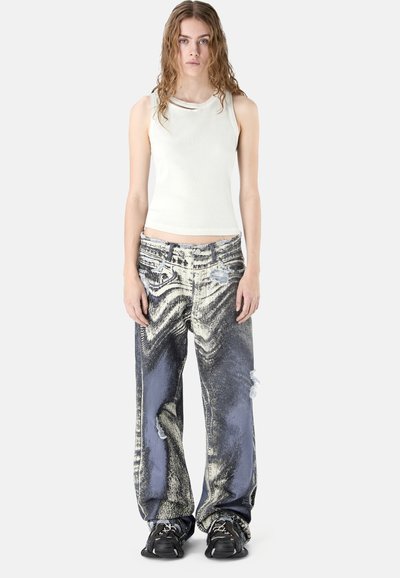 Jeune personne aux cheveux ondulés portant un débardeur blanc, un jean large délavé à l'acide avec des déchirures, et des baskets noires, debout devant un fond uni.