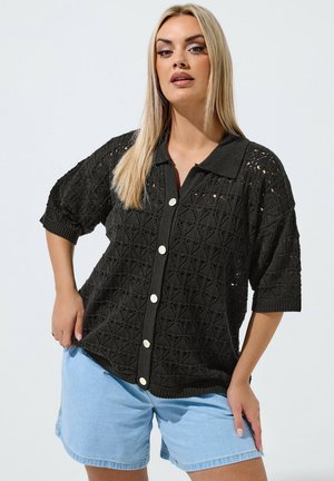 Lyshåret kvinde iført en sort kortærmet strikket knap-up skjorte og lyseblå denimshorts, stående med den ene hånd på hoften.
