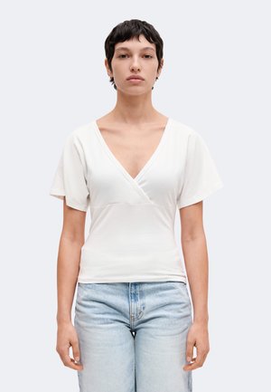 Jeune personne aux cheveux courts et foncés, portant un haut blanc à manches courtes en V et un jean bleu clair, faisant face à un fond uni.