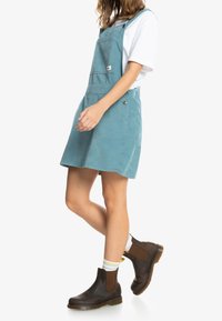 Tyrkysové bavlněné šaty overal s nastavitelnými ramínky, přední kapsou a rozšířeným lemem. V kombinaci s hnědými chelsea botami a bílými ponožkami.