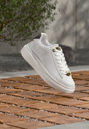 Baskets en cuir blanc avec des accents dorés et un talon marron texturé, flottant au-dessus d'un sol en carreaux de bois avec de petits cailloux entre les carreaux à l'extérieur.