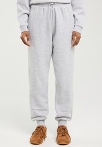 Osoba ubrana w jasnoszare spodnie dresowe i brązowe sznurowane trampki, stojąca na jednolitym, jasnym tle.