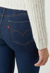 Persona vestida con jeans oscuros de Levi's y un suéter de punto blanco, con la mano relajada a su lado contra un fondo sencillo.