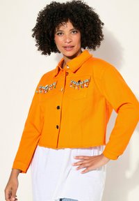 Orange denim kort jacka med svarta knappar, med broderade mönster och färgglada detaljer på bröstfickorna.