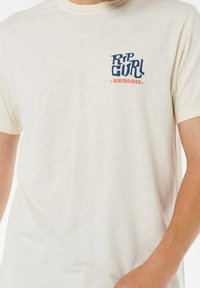 Kremowa bawełniana koszulka z krótkim rękawem, z niebiesko-czerwoną logo "Rip Curl Surfboards" na lewej klatce piersiowej.