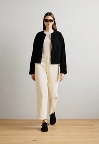 Jacket scurt texturat cu blana neagră, cu fermoar în față, purtat peste un top alb cu dungi, asociat cu pantaloni crem cu talie înaltă și pantofi negri.