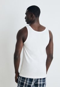 Top sin mangas de algodón blanco con un escote redondo, que cuenta con una textura suave. La persona que lo lleva puesto está combinada con pantalones cortos de cuadros, mostrando un ajuste relajado.