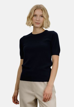 En kvinne med blondt hår, iført en svart kortermet genser og beige bukser, står mot en ensfarget lys bakgrunn.