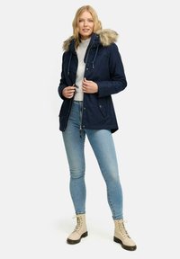 Navy parka jacket with faux fur hood, drawstring, and zipper closure. Paired with light blue jeans and beige lace-up boots.