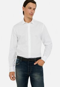Camicia bianca a maniche lunghe con colletto, chiusura a bottoni e texture liscia, abbinata a jeans in denim blu scuro e una cintura nera.