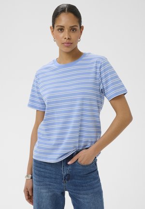 Femme portant une chemise à manches courtes rayée bleu clair et blanc et un jean bleu, debout avec une main dans la poche devant un fond uni.