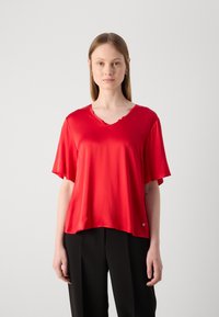 Blusa rossa a maniche corte realizzata in tessuto leggero con scollo a V e orlo ondulato. Presenta un piccolo logo accento vicino in basso a sinistra.